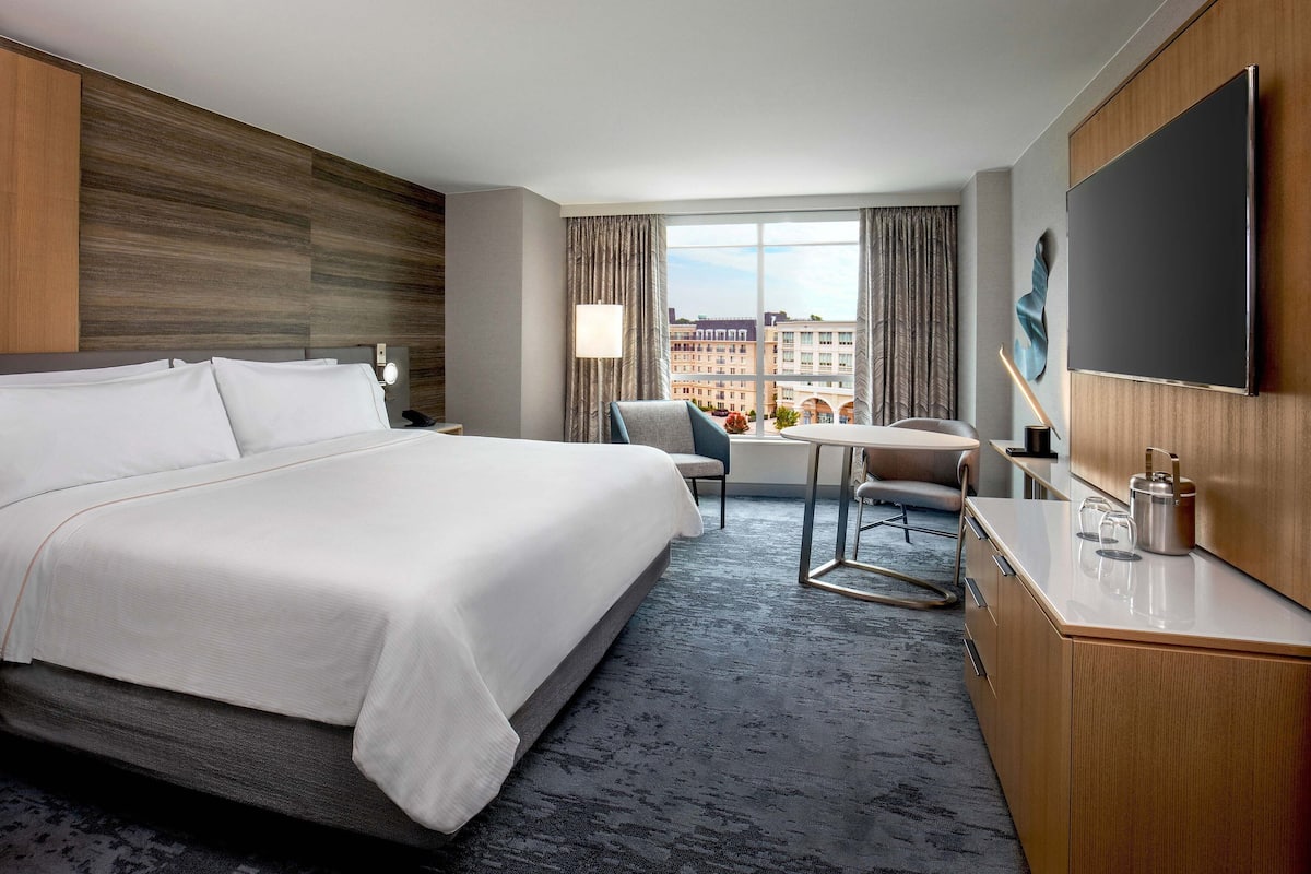 A hotel room with a large bed, a desk, a chair, a TV, and a view of the city.