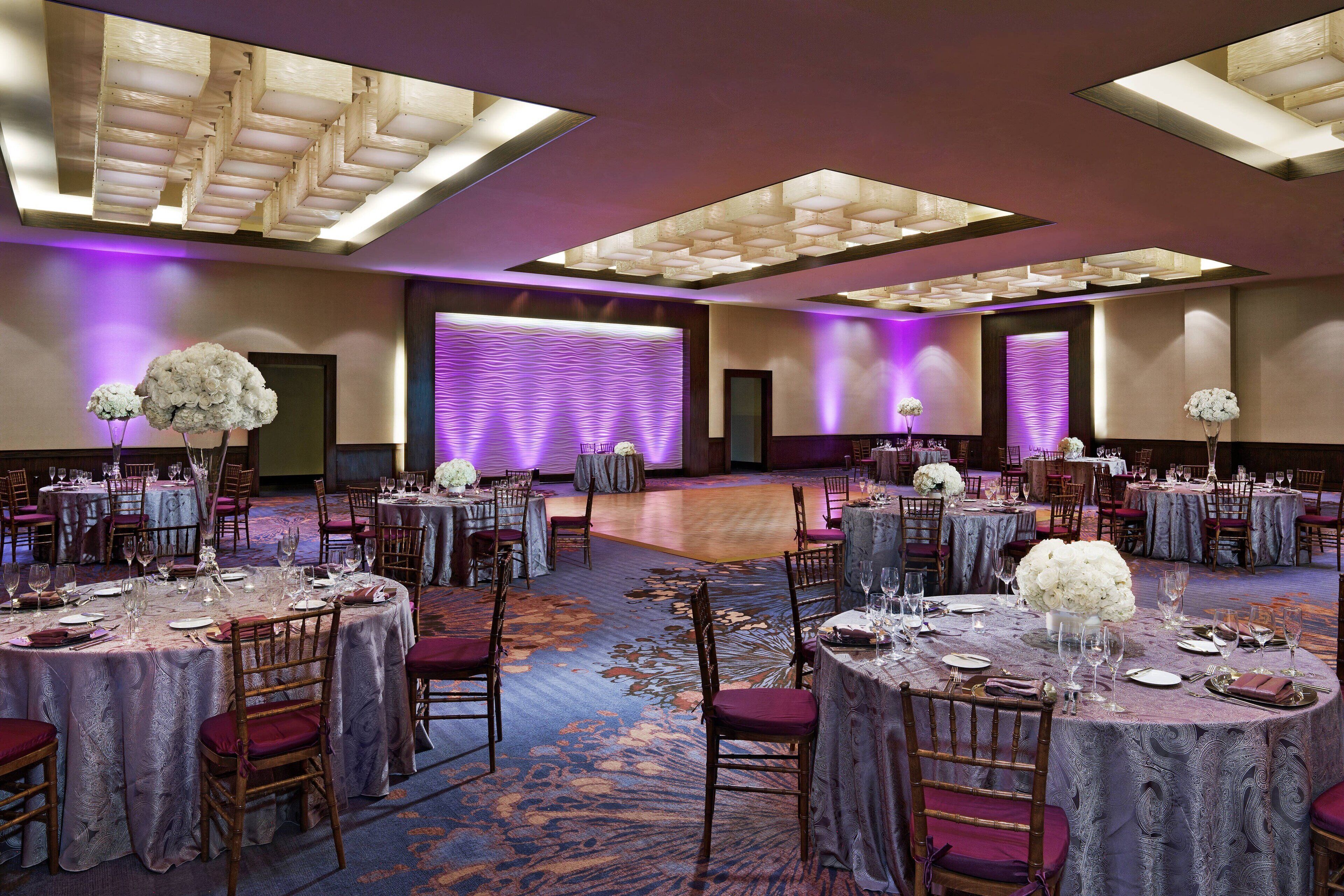 A banquet hall with round tables, chairs, and floral centerpieces, featuring a patterned carpet and modern lighting fixtures.