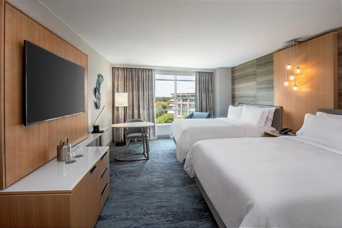 A hotel room with two beds, a flat-screen TV, a desk with a lamp, and a view of buildings through the window.