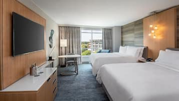 A hotel room with two beds, a flat-screen TV, a desk with a lamp, and a view of buildings through the window.