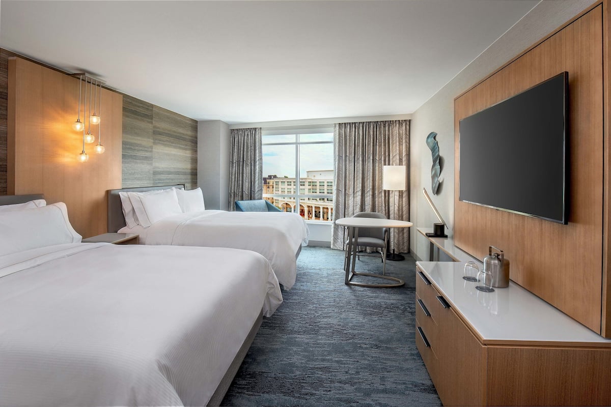 A hotel room with two beds, a desk, a flat-screen TV, and a view of the city.