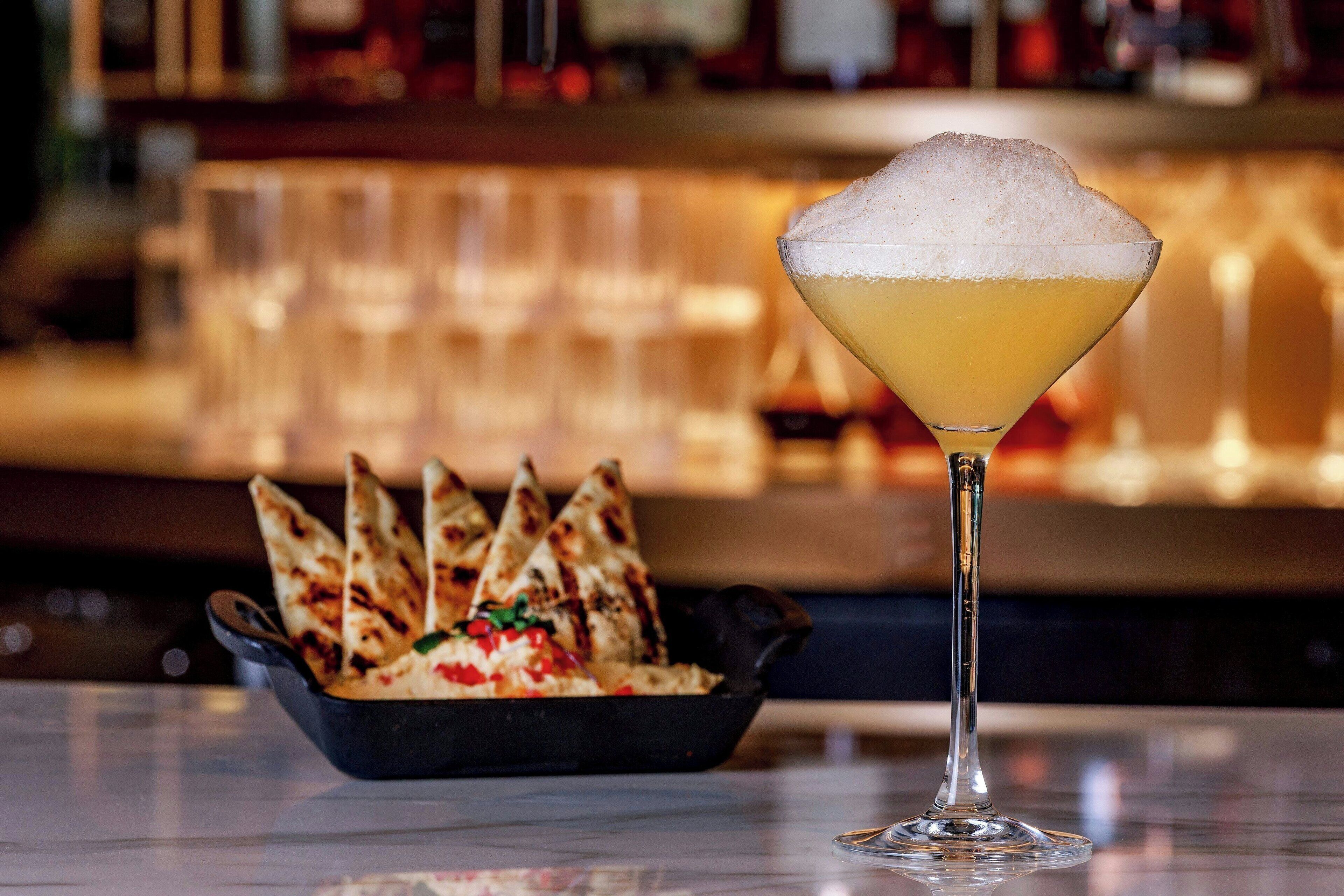 A martini glass with a frothy top and a bowl of grilled flatbread with toppings.