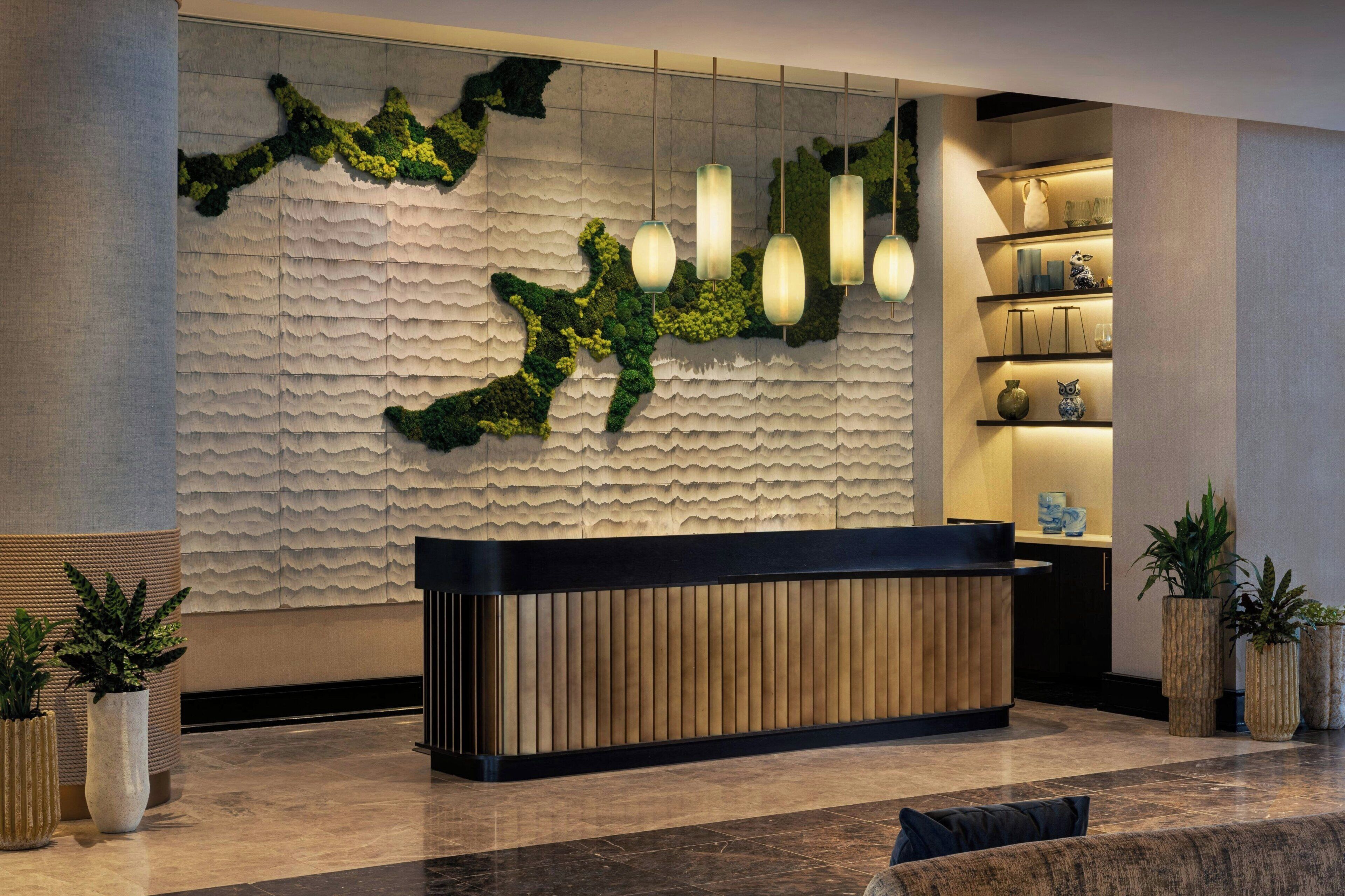 A reception area with a textured wall, a wooden reception desk, hanging pendant lights, and potted plants.