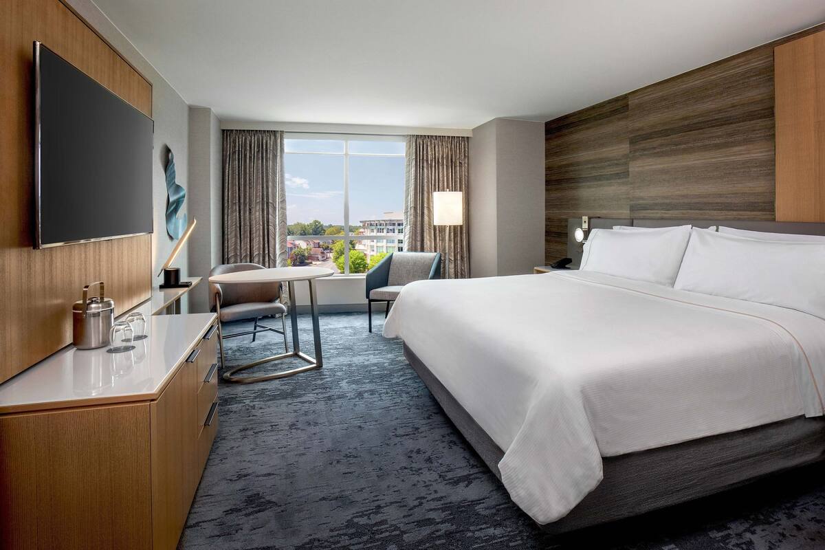 A hotel room with a large bed, a desk with a flat-screen TV, a chair, and a view of the city through large windows.