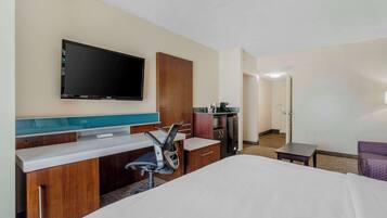 A hotel room with a bed, a desk with a chair, a television mounted on the wall, a kitchenette, and a door leading to another room.