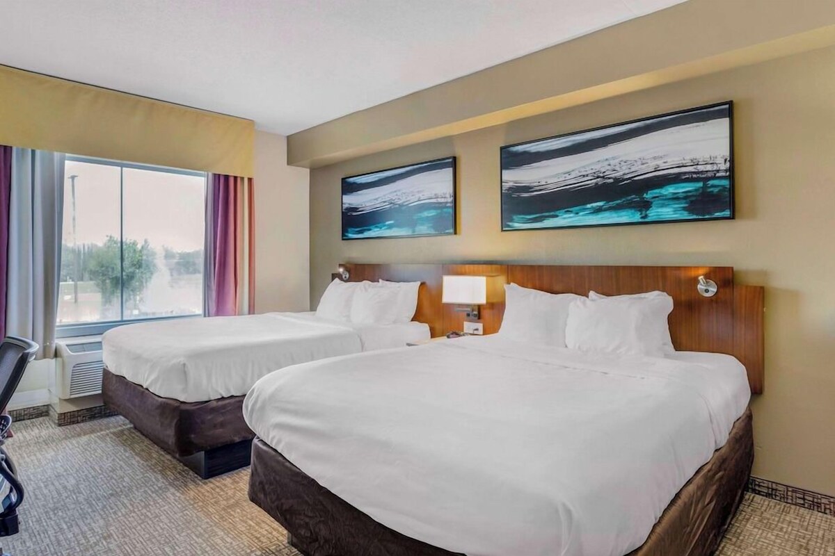 A hotel room with two beds, a desk, a chair, and large windows.