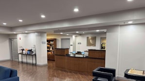 A hotel lobby with a reception desk, a blue sofa, a chess table, and a kitchen area in the background.