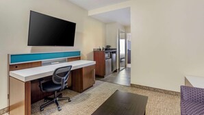 A hotel room with a desk, chair, flat-screen TV, kitchenette, and a view of the hallway.