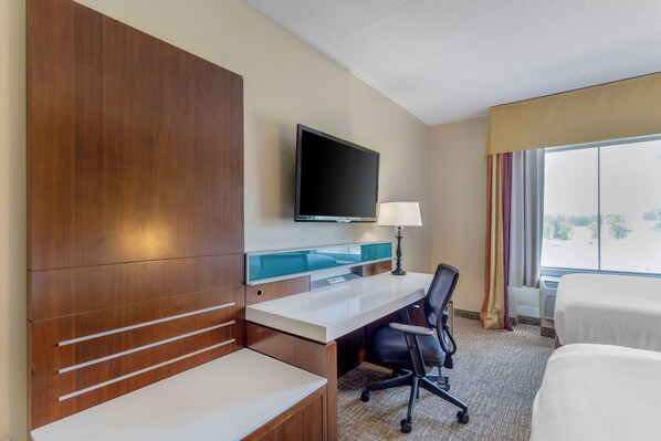 Habitación de hotel con escritorio de madera, televisor de pantalla plana, lámpara, una silla y dos camas.