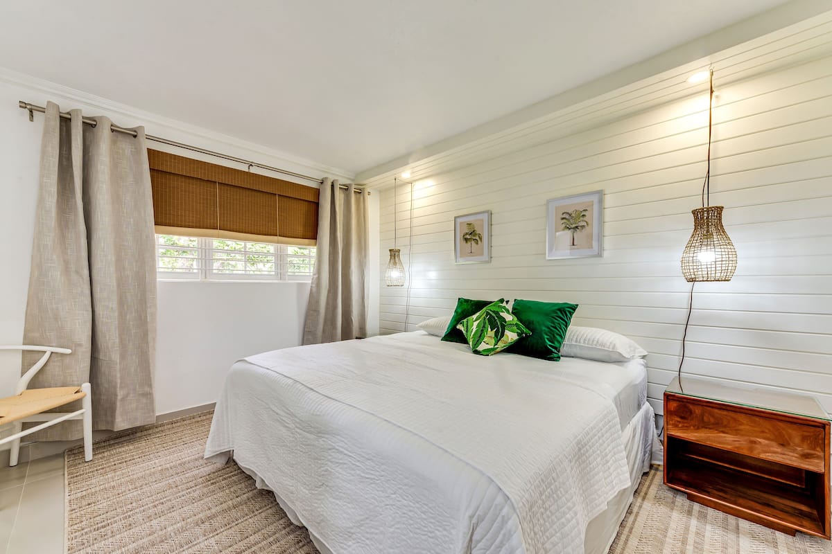 A bedroom with a bed, a bedside table, a chair, a window with curtains, and wall-mounted lights.