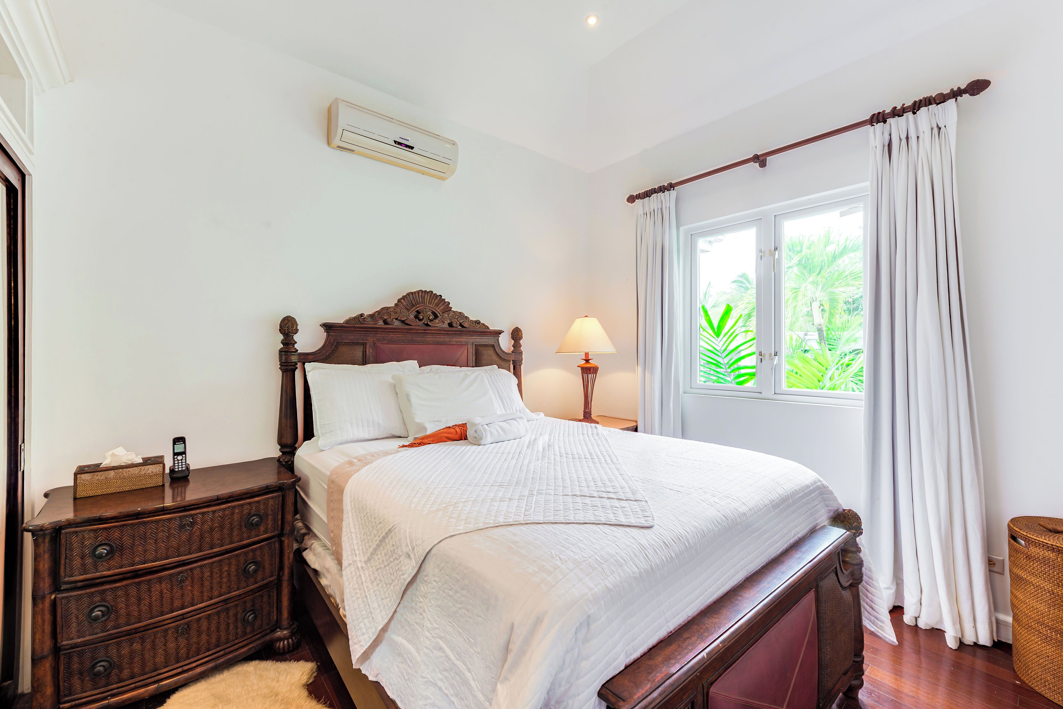 A neatly made bed with a wooden headboard, a bedside table with a lamp, and a window with curtains.