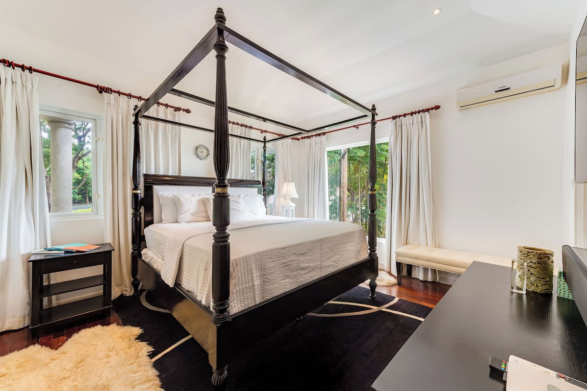 A four-poster bed with a white bedspread, a bedside table with a lamp, and a view of greenery outside.