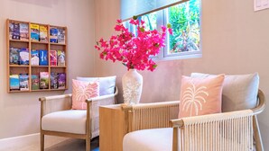 A cozy seating area with wicker chairs and a white vase with pink flowers. A wooden bookshelf with travel books is visible in the background.