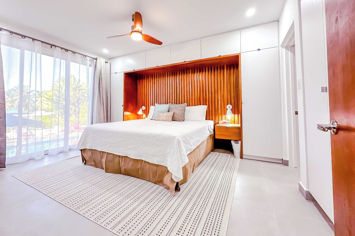 A modern bedroom with a large bed, a ceiling fan, and a view of the outdoors through the window.