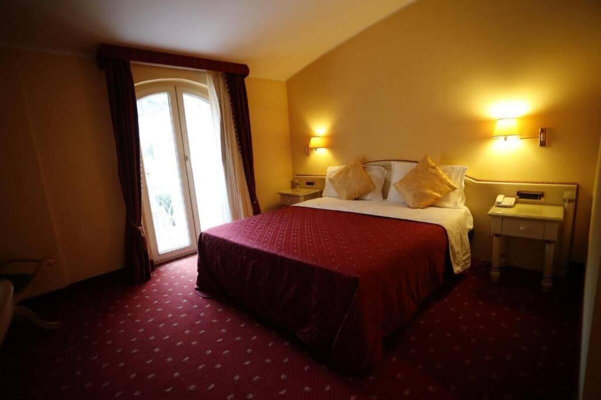 A hotel room with a large bed, two bedside tables, a window with curtains, and wall-mounted lights.