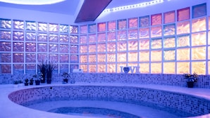 Uma área de spa com uma banheira de hidromassagem com mosaicos, plantas em vasos e uma parede de janelas com vidro jateado.