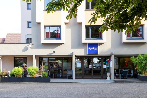 A hotel with a sign reading 'Kyriad Hotel' above the entrance, featuring a modern architectural style with large windows and a flat roof.