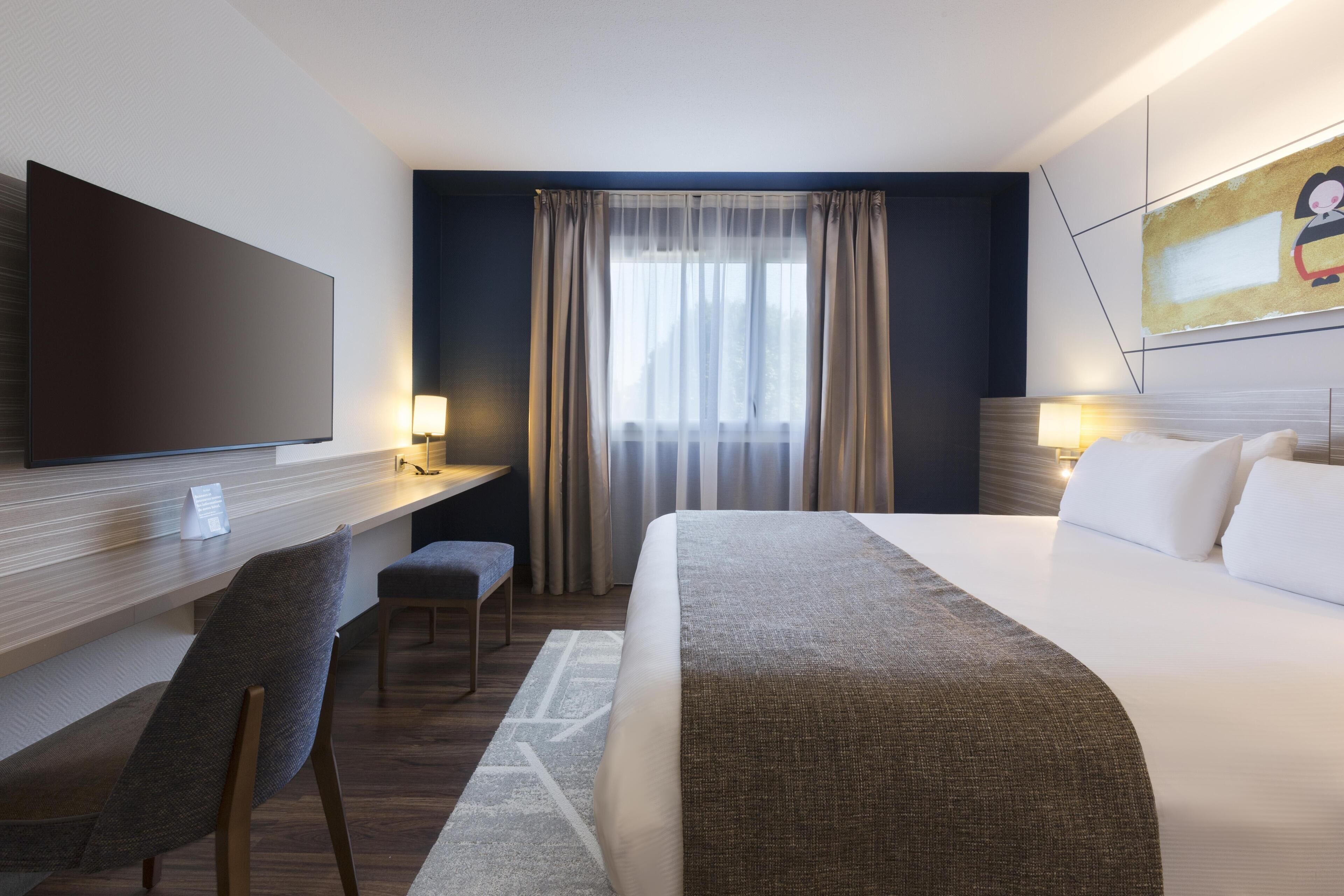 Une chambre d’hôtel avec un lit, un bureau, une chaise, une télévision et une fenêtre avec des rideaux.