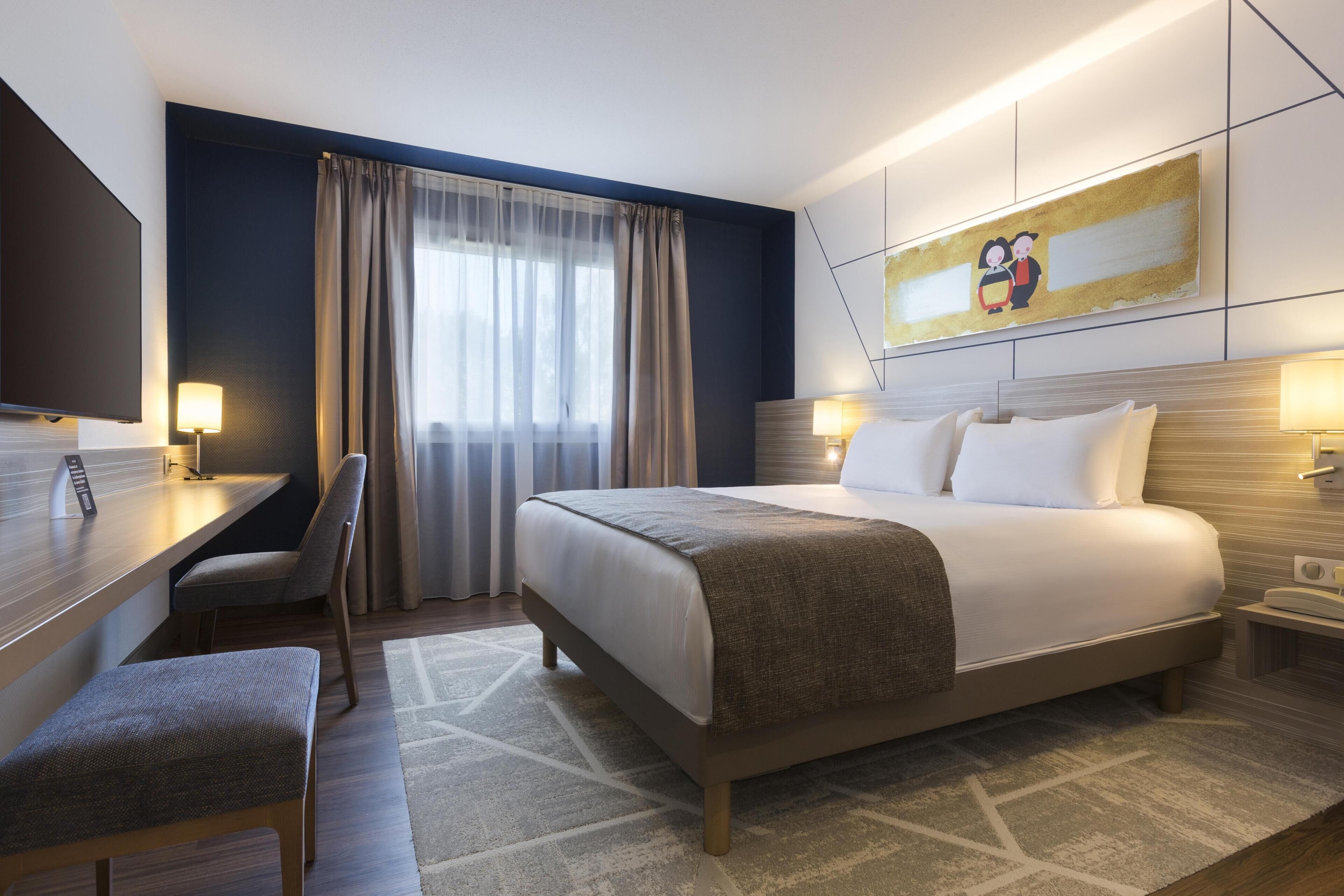 A hotel room with a bed, bedside table, chair, desk, and a TV.
