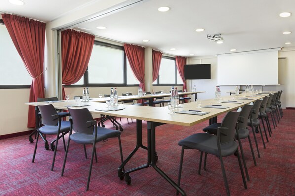 A conference room with a long table, chairs, a projector screen, and windows with curtains.