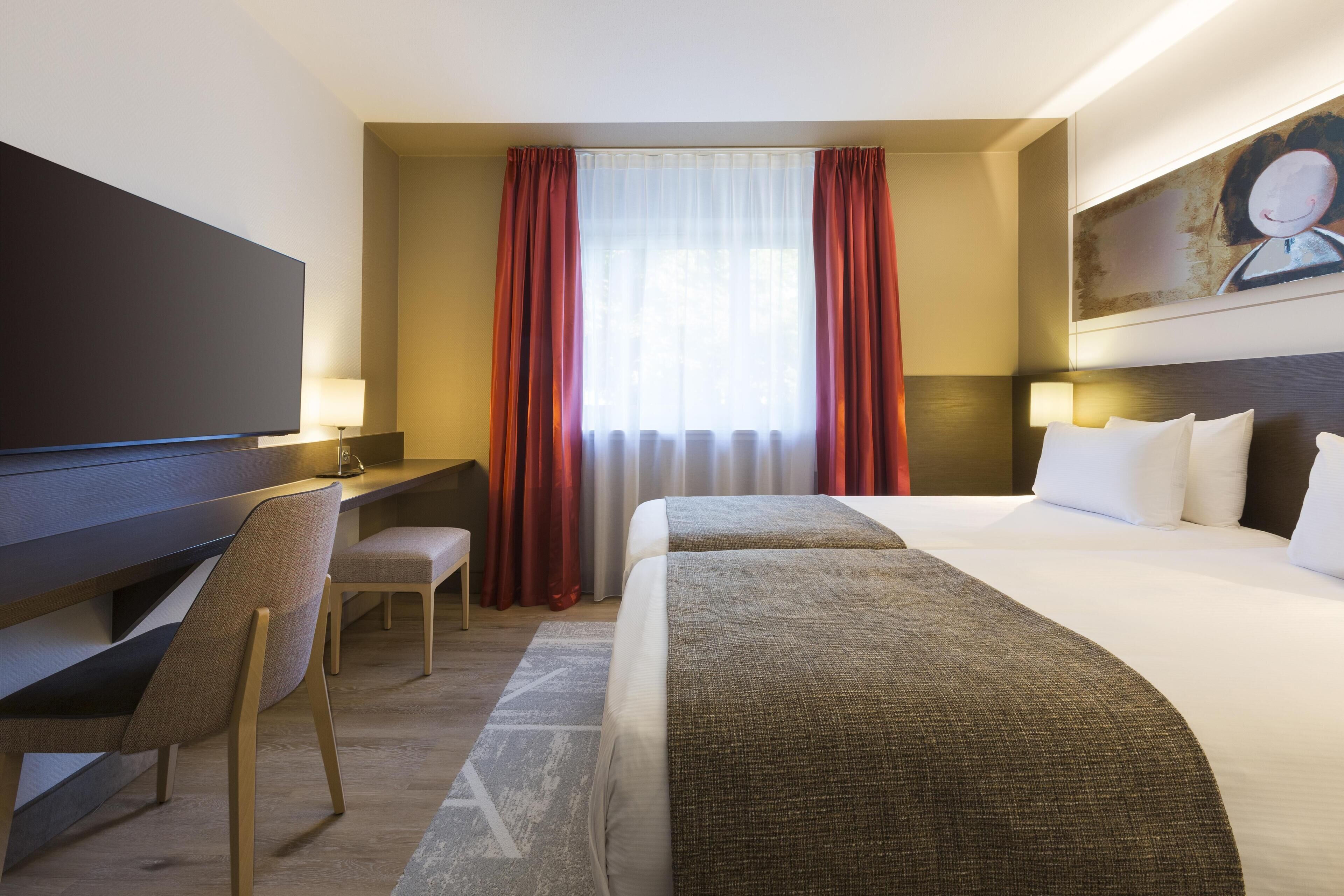 A hotel room with a bed, a desk, a chair, a TV, and a painting on the wall.