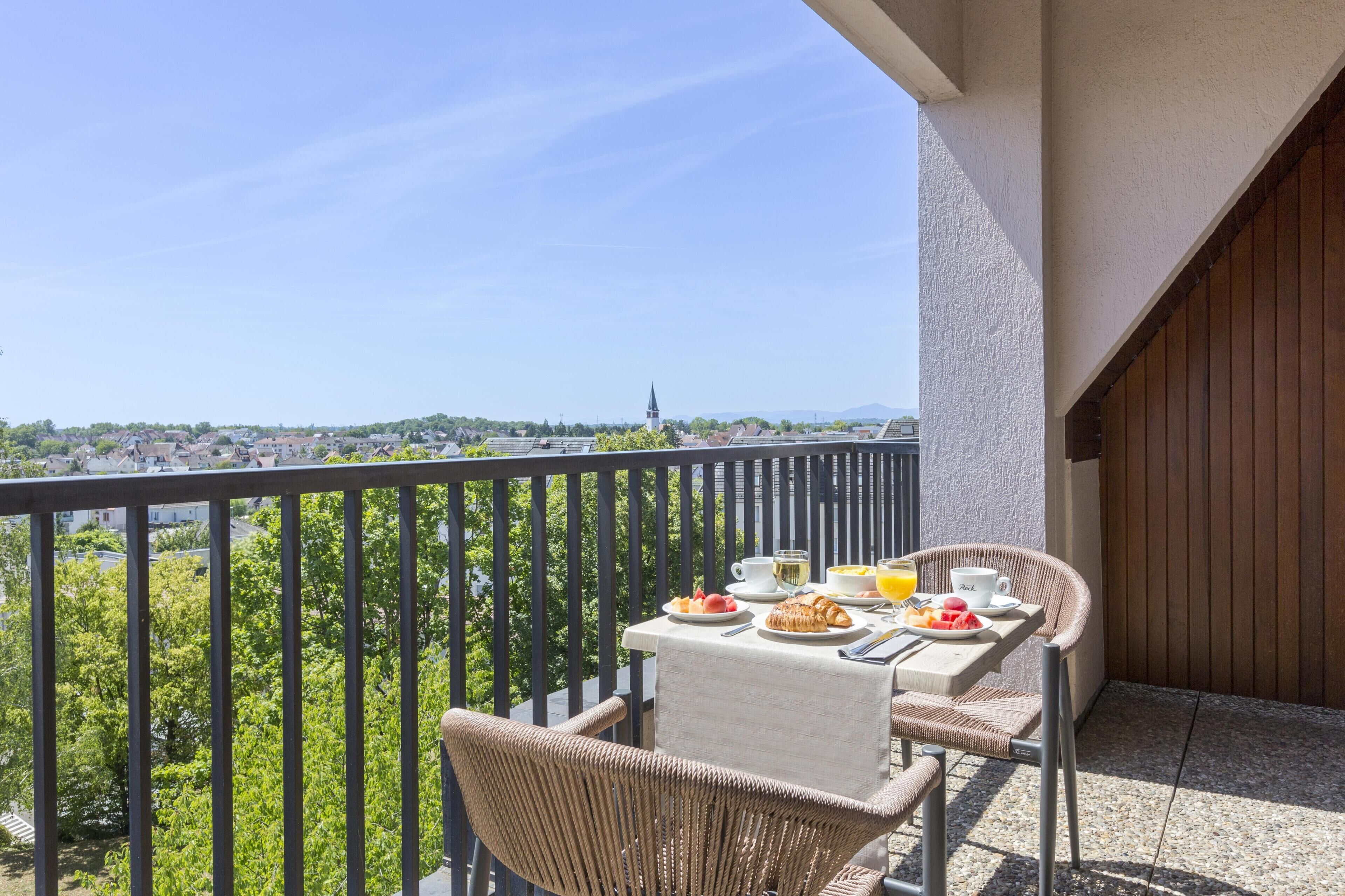 Balcón con una mesa dispuesta para el desayuno, con vistas a una zona residencial con casas y la aguja de una iglesia.