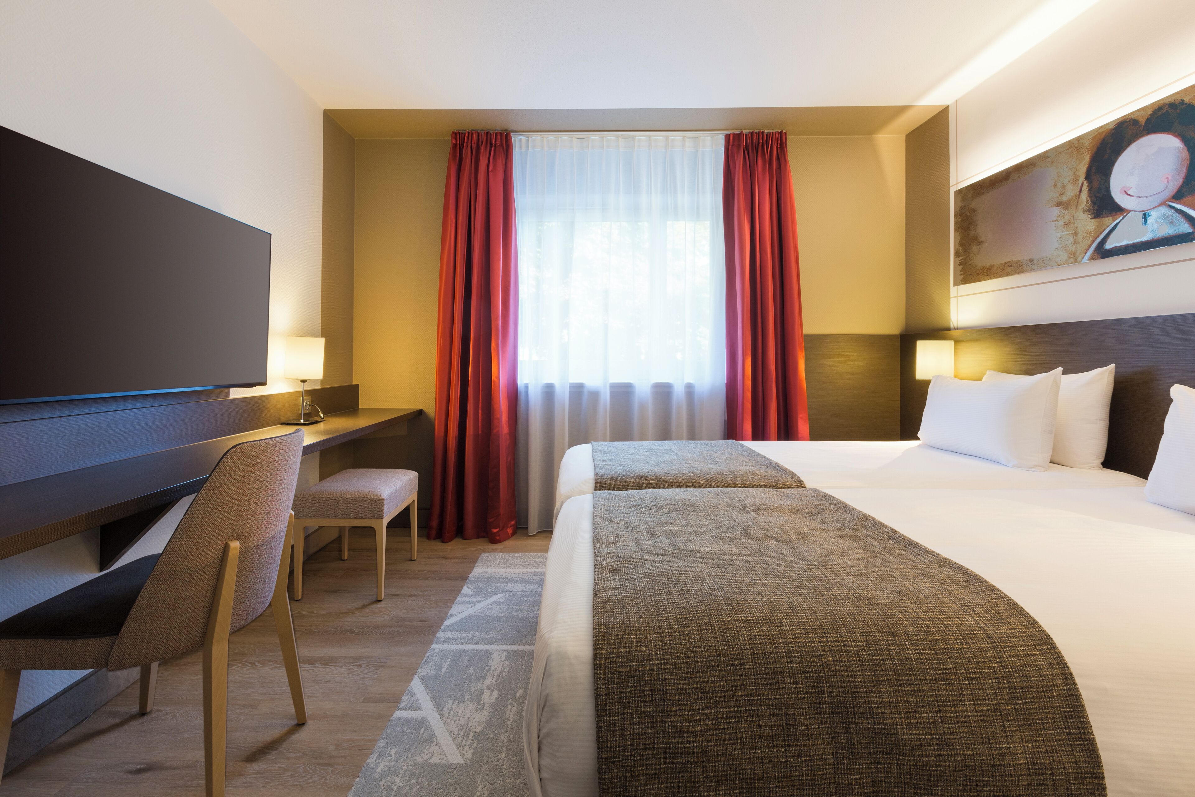 Quarto de hotel com cama, escrivaninha, cadeira, televisão e um quadro na parede.