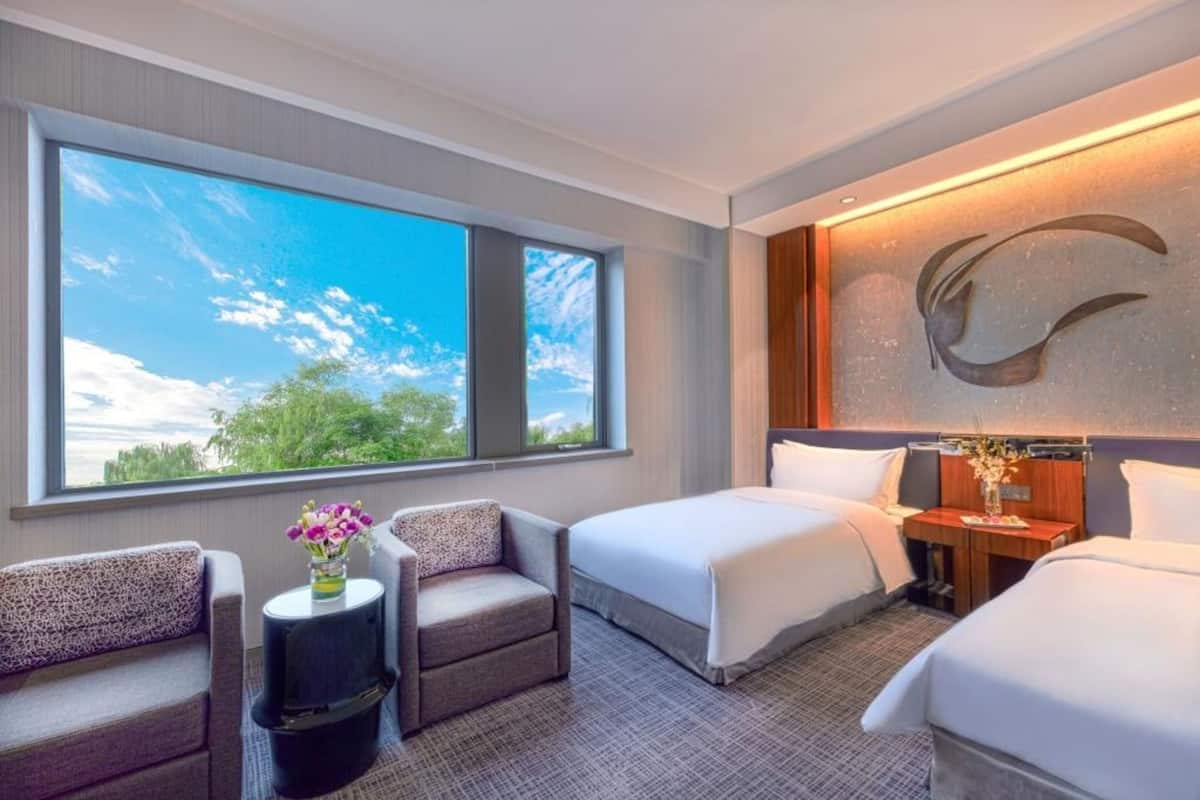 A hotel room with two beds, a sofa, a small table with a vase of flowers, and a large window offering a view of trees and sky.