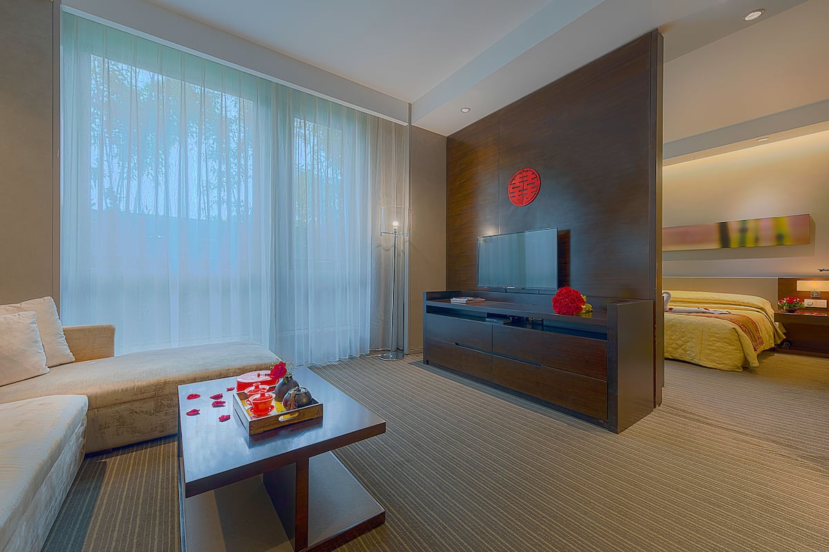 A modern hotel room with a sofa, a coffee table with rose petals, a TV, and a bed with a yellow bedspread.