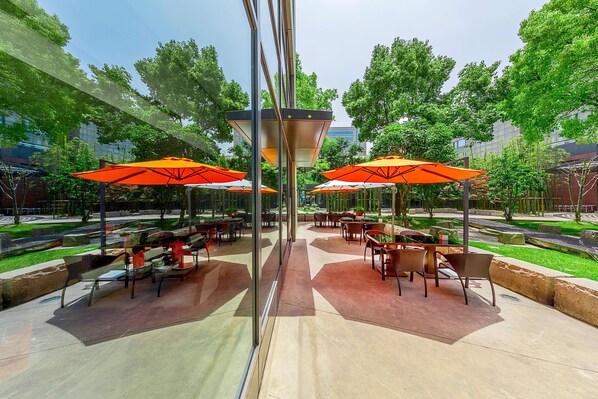 An outdoor patio area with tables and chairs under orange umbrellas, surrounded by greenery and a clear sky. - Shanghai Zhangjiang Parkyard Hotel (Shanghai)