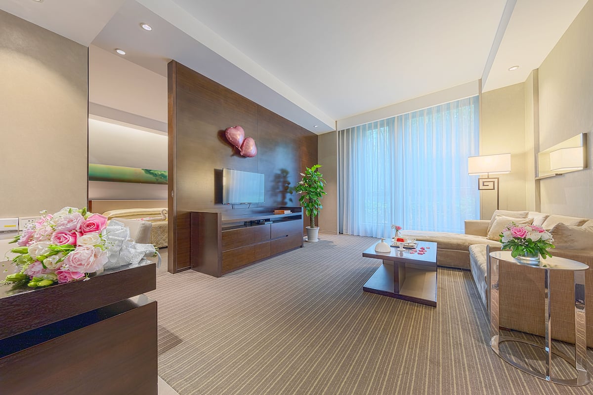 A modern hotel room with a sofa, a coffee table, a TV, a plant, and a desk with flowers.