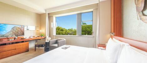 A hotel room with a large bed, a desk, a chair, a television, and a window with a view of greenery.