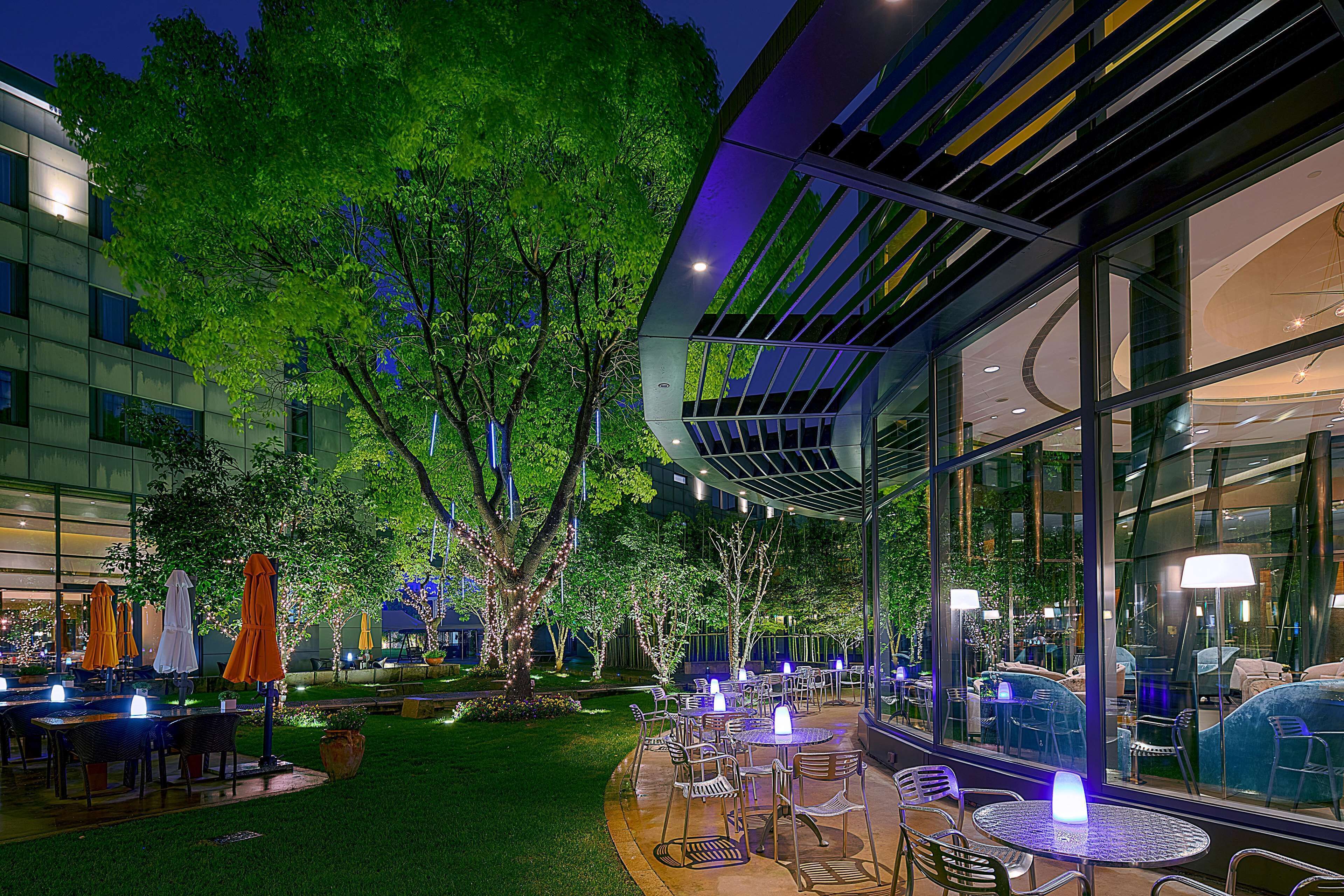 An outdoor dining area with tables and chairs, surrounded by trees and a building with large windows.