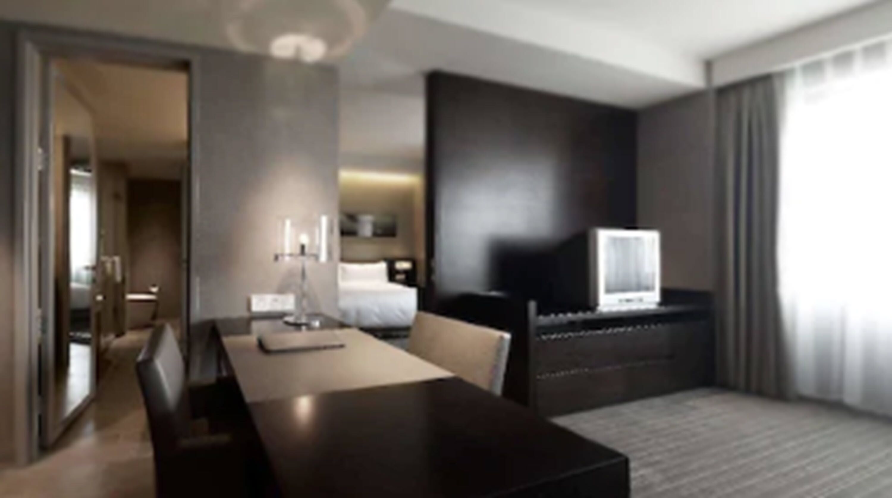 Una habitación de hotel moderna con una cama de tonos oscuros, un televisor de pantalla plana sobre un mueble, una mesa de comedor con sillas y un amplio ventanal con cortinas.