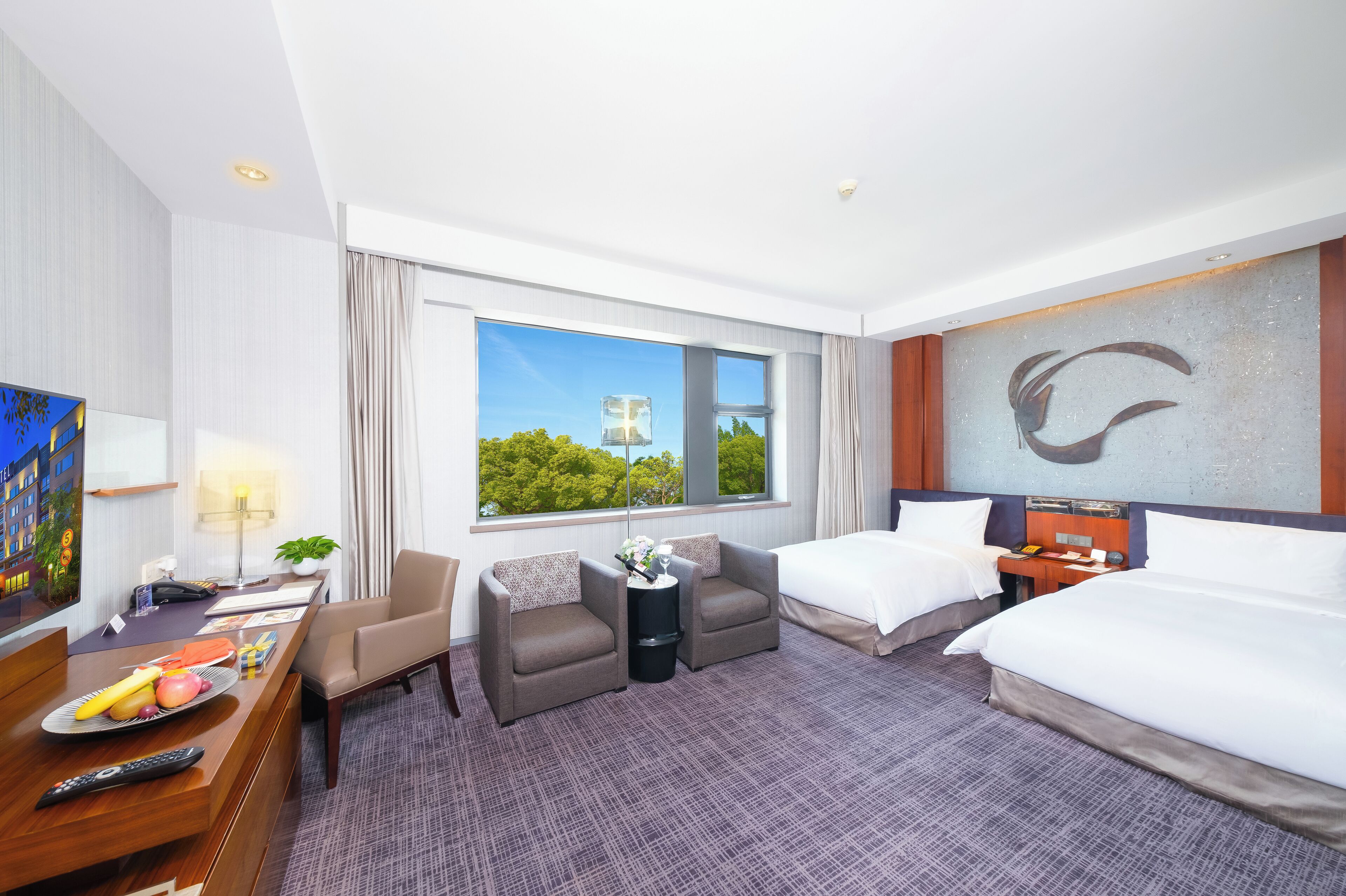 A hotel room with two beds, a desk with a computer, a chair, a small table with a plant, and a large window with a view of trees and a building.