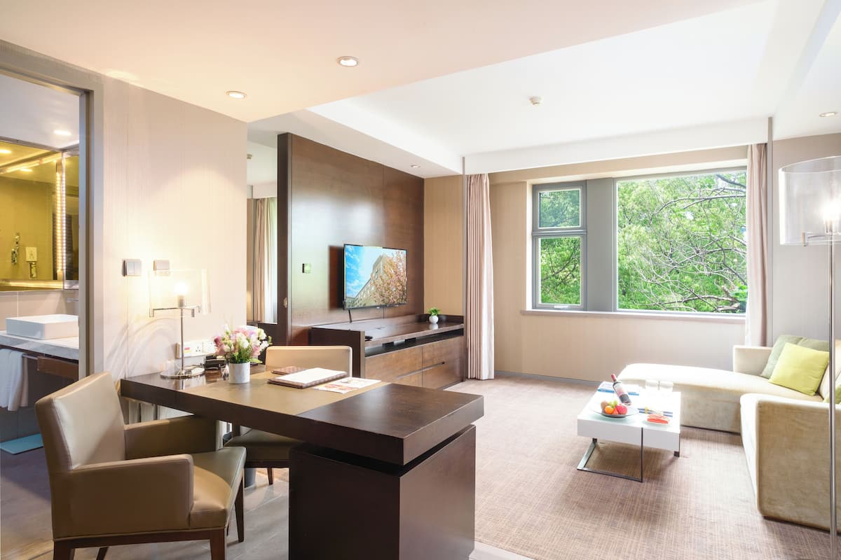 A modern hotel room with a dining area, a sofa, a TV, and a large window with a view of trees.