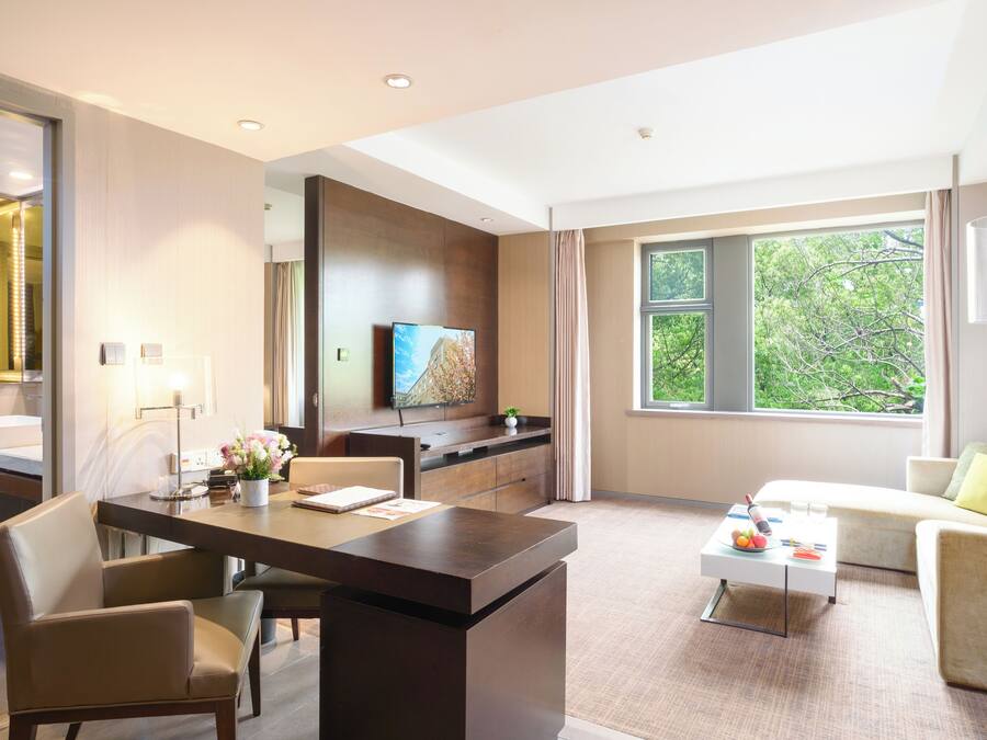 Quarto de hotel moderno com área de refeições, sofá, televisão e uma grande janela com vista para árvores.