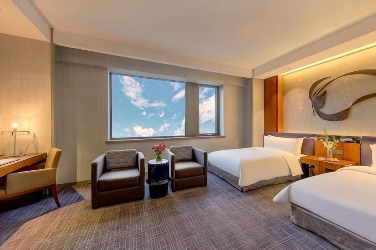 A hotel room with two beds, a desk, a chair, a small table with a vase, and a large window.