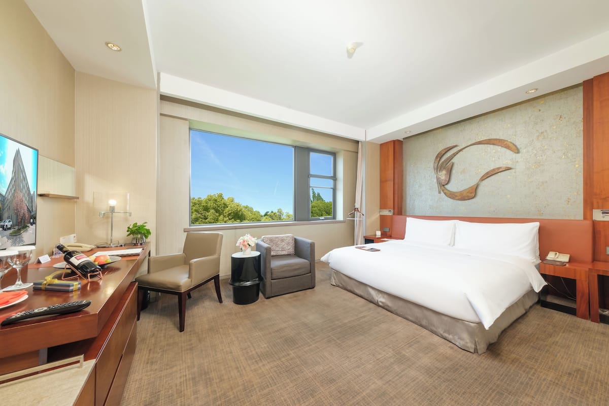 A modern hotel room with a large bed, a desk with a chair, a small sofa, and a large window offering a view of greenery.