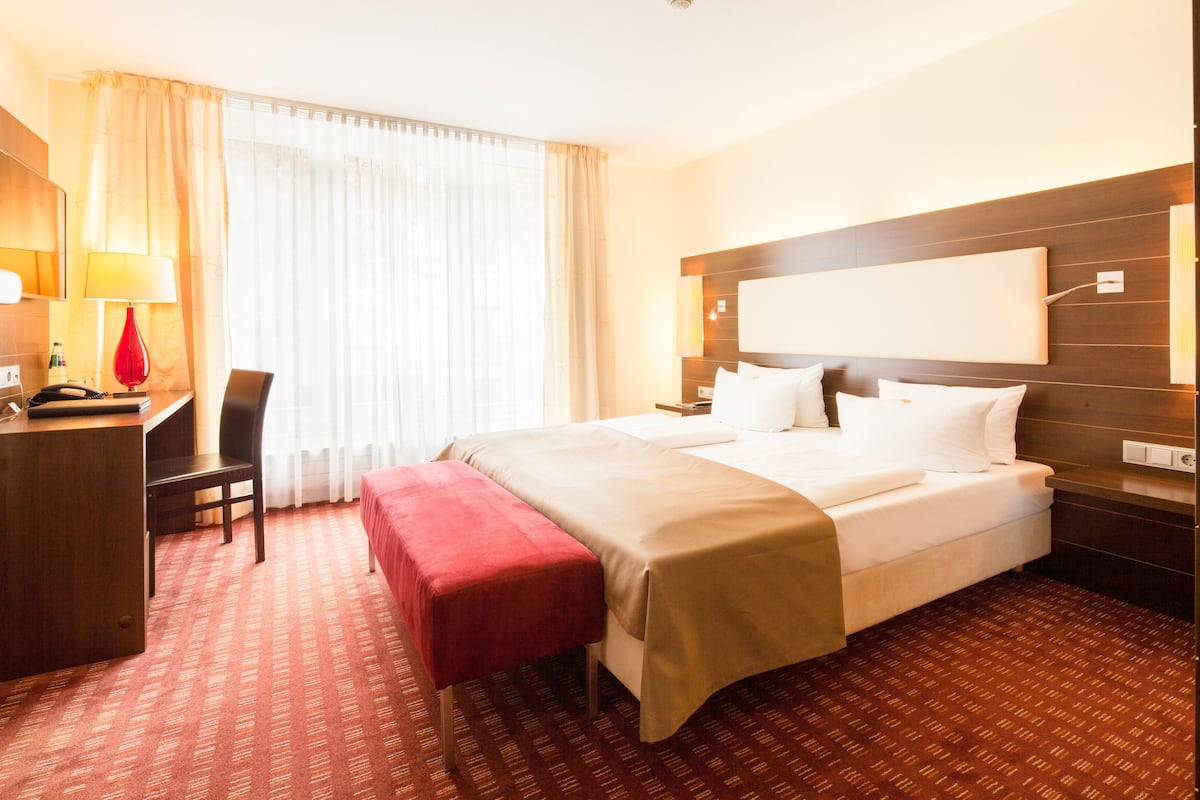 Hotelkamer met een bed, een bureau, een stoel, een rode bank en een groot raam met gordijnen.