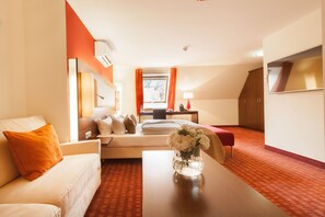 Habitación de hotel con cama, sofá, mesa con un jarrón de flores y televisión en la pared.