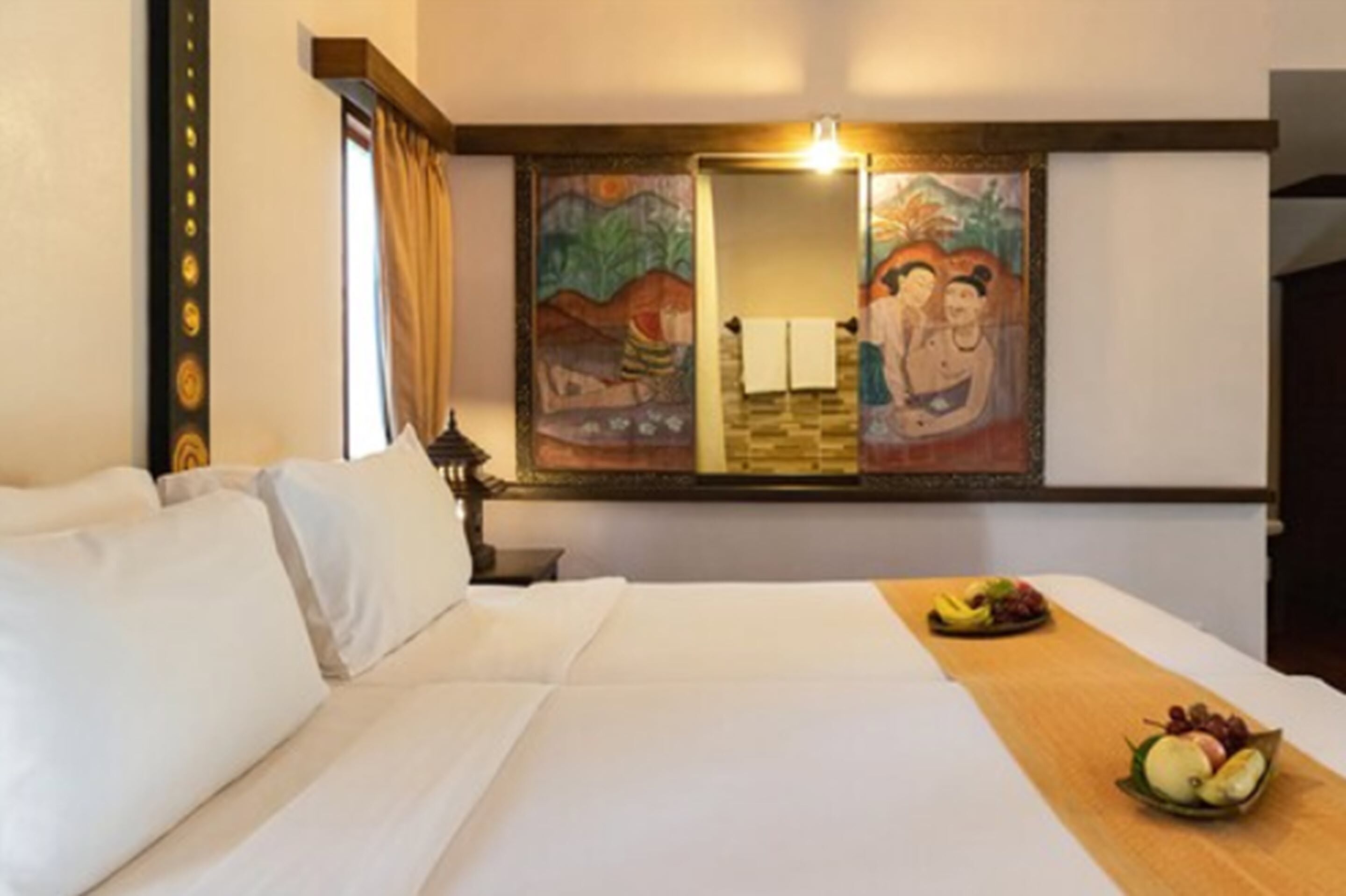A hotel room with a bed, a wooden side table with fruit, a painting on the wall, and a mirror.