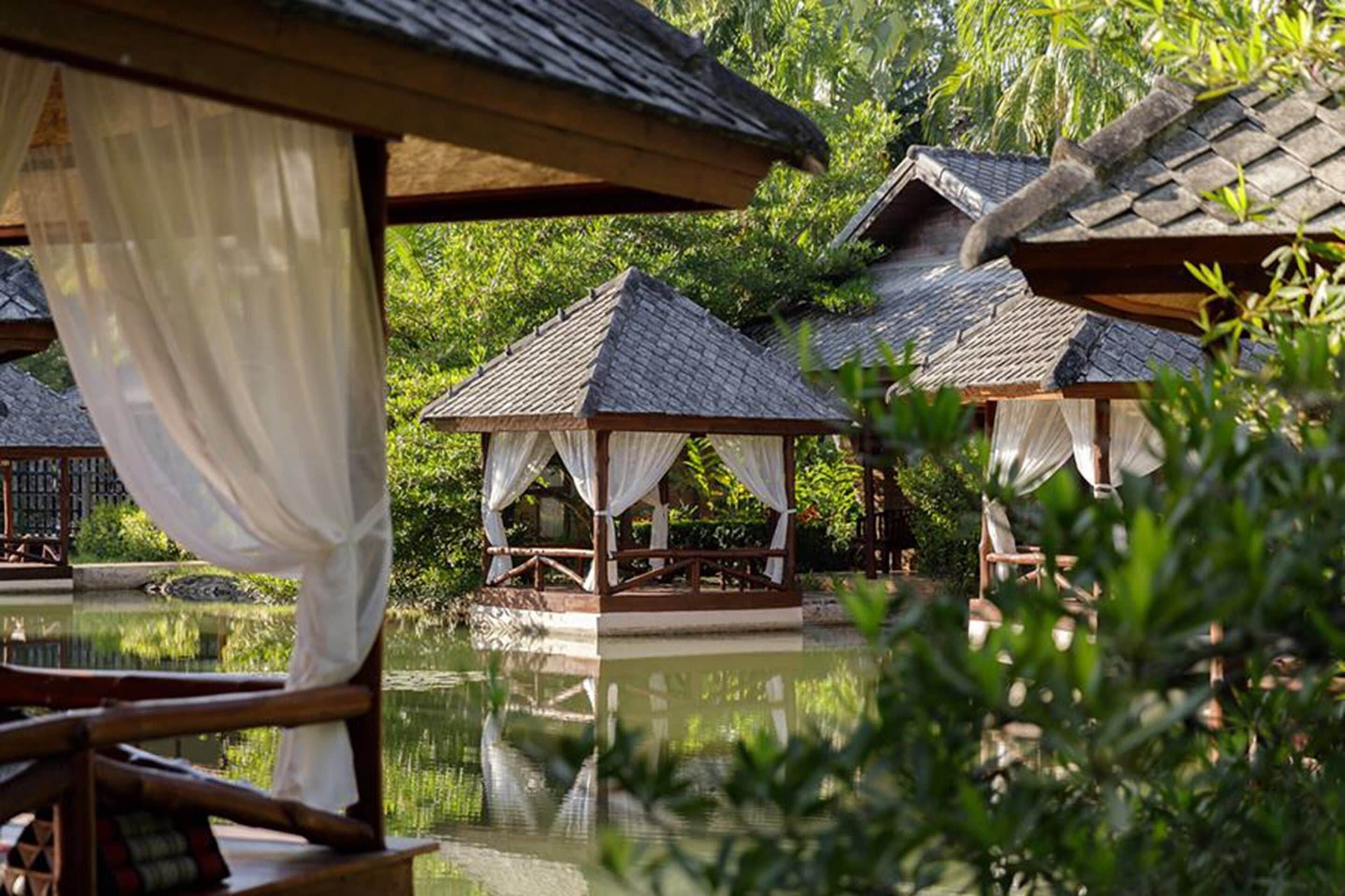 A tranquil outdoor setting with thatched-roof structures, a reflective water body, and lush greenery.