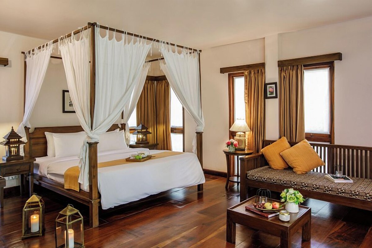 A four-poster bed with a canopy, a sofa with cushions, a bedside table with a lamp, and a tray with fruits and a vase.