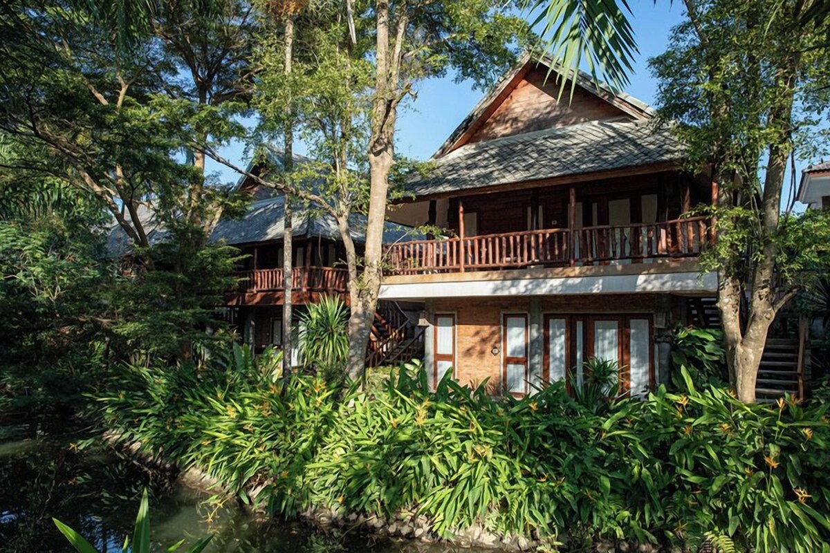 A traditional wooden house with a balcony, surrounded by lush greenery and a small pond.