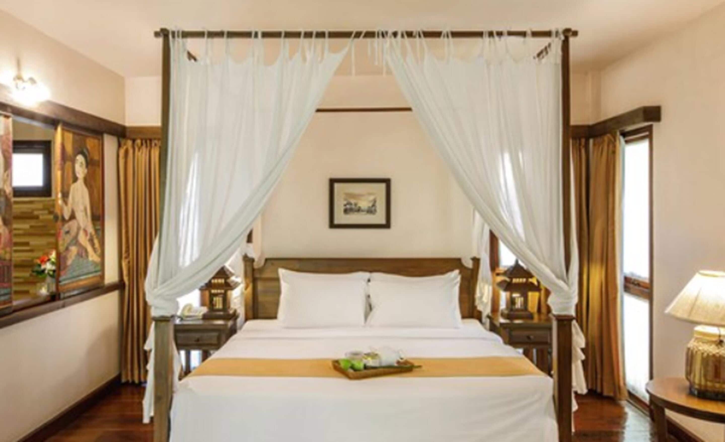 A hotel room with a four-poster bed, white curtains, wooden headboard, and a small table with a tray of food.