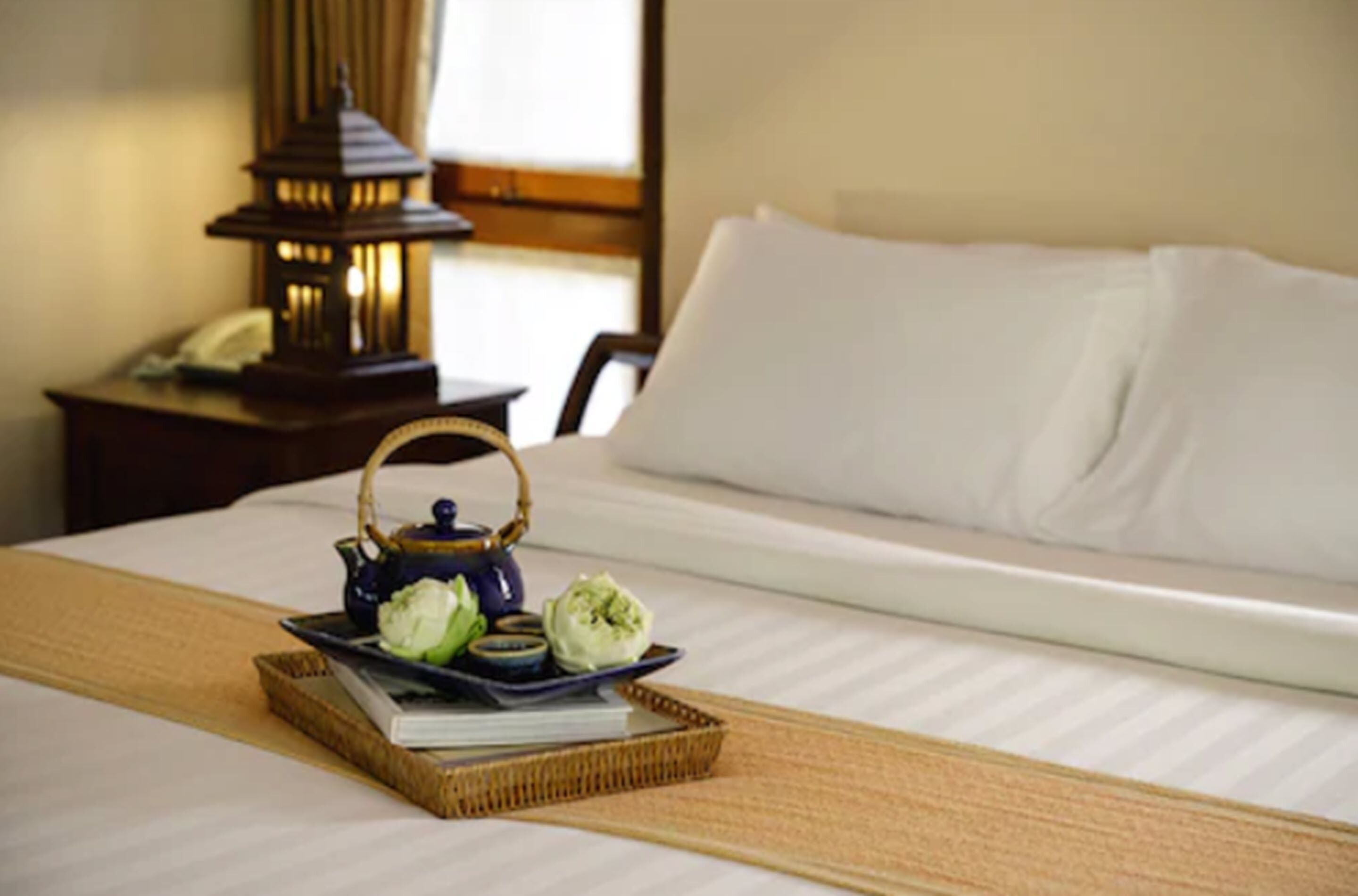 Una cama hecha con esmero, una bandeja con té y aperitivos, una lámpara y una mesita de noche con un teléfono.