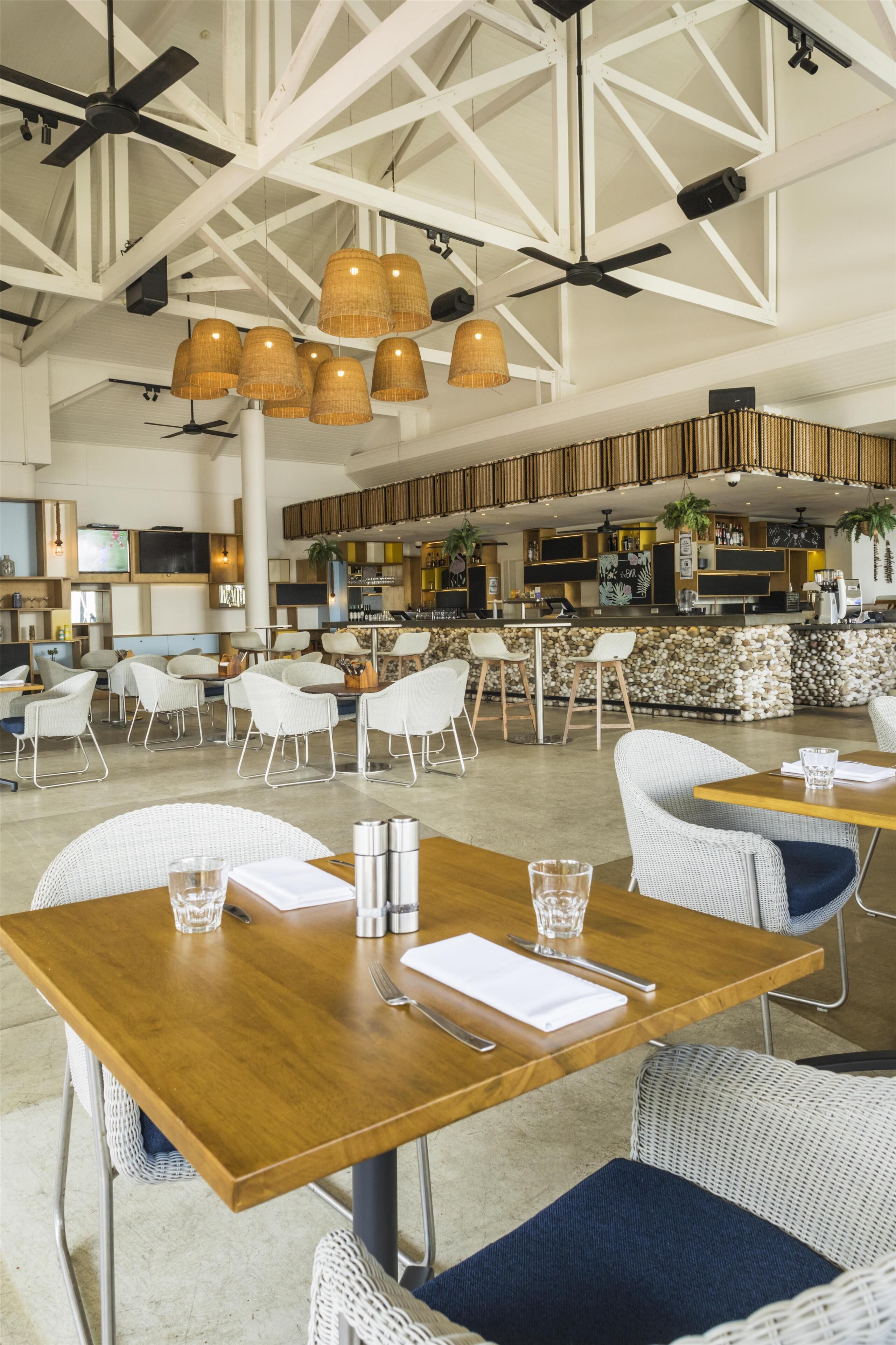 A spacious restaurant with high ceilings, exposed beams, and hanging wicker light fixtures. Tables are set with white chairs and have water glasses and condiments.