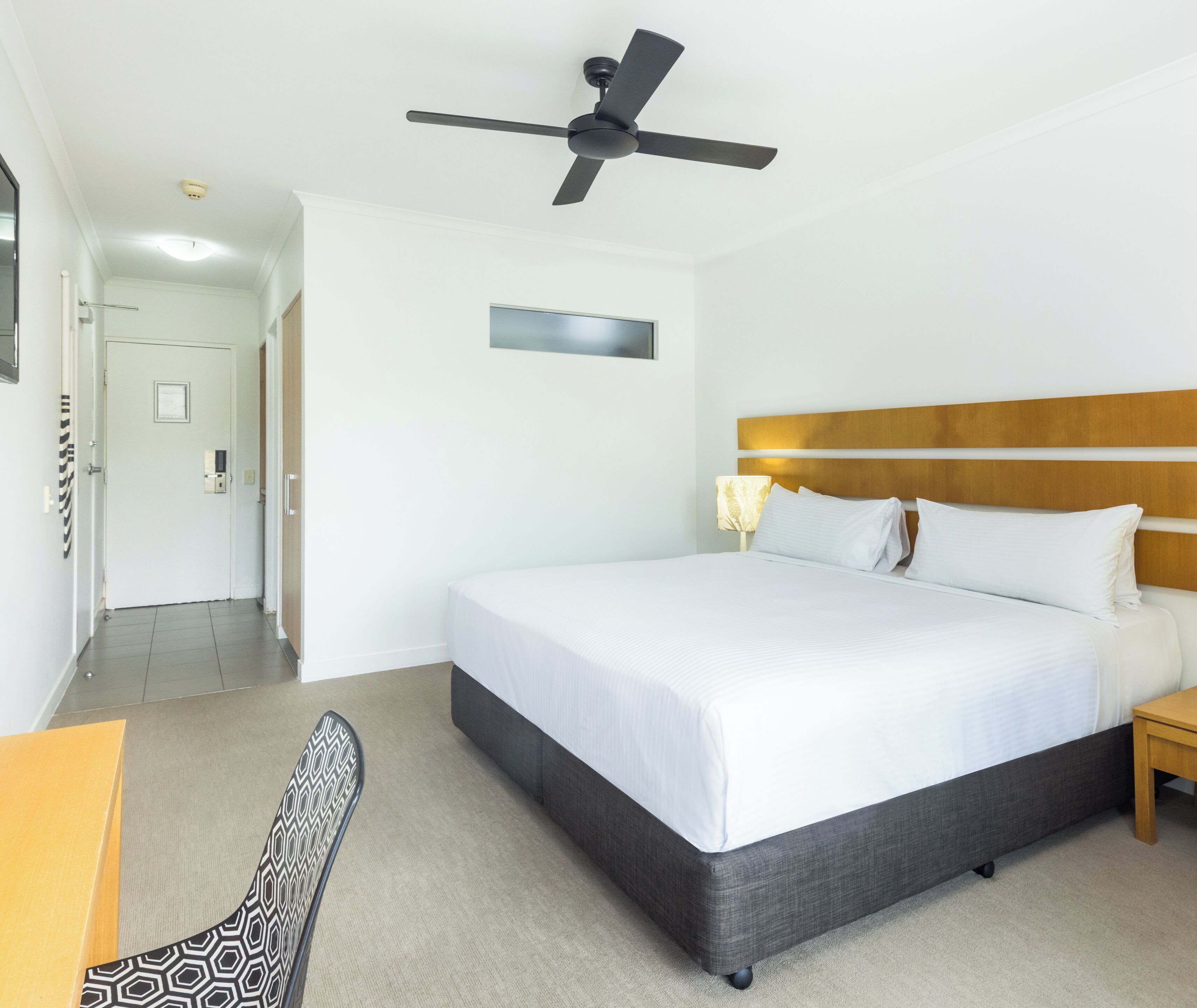 A hotel room with a bed, a ceiling fan, a nightstand, a lamp, and a chair.