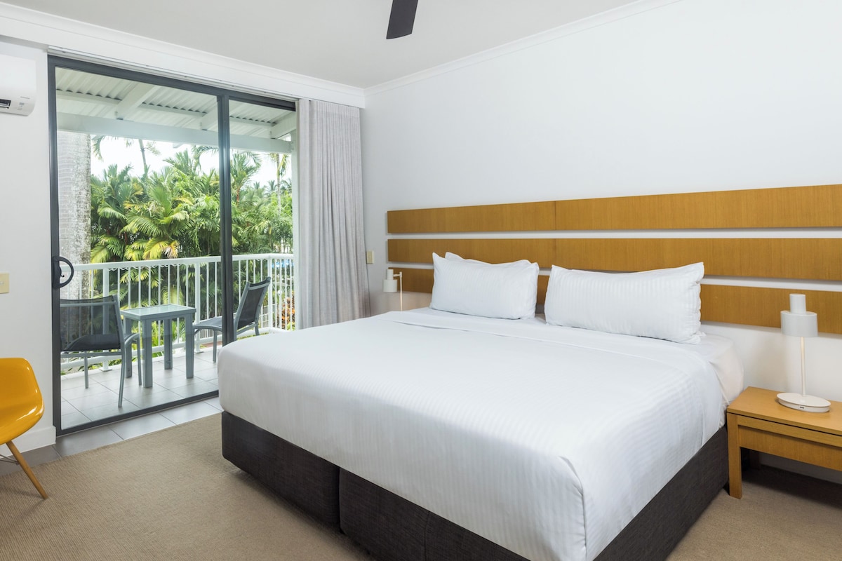 A hotel room with a large bed, a ceiling fan, a nightstand, a small table, a chair, and a balcony with a view of palm trees.